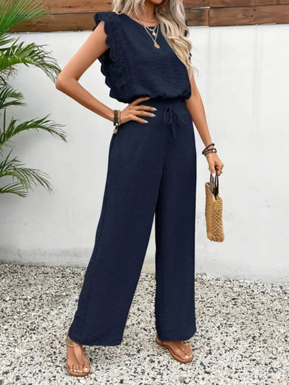 Woman wearing a navy blue jumpsuit with a white wall and plant in the background